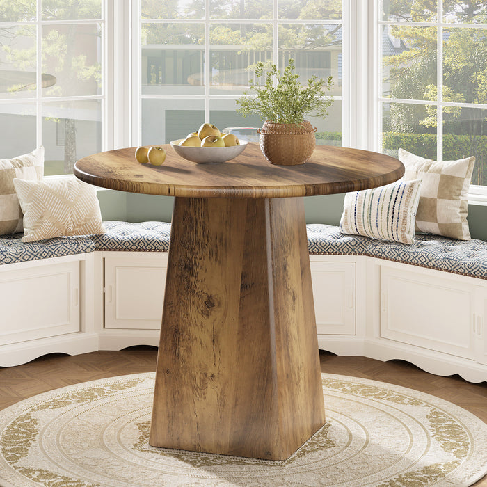 Wooden dining table with a round top and a single pedestal base in a room with large windows.