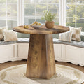 Wooden dining table with a round top and a single pedestal base in a room with large windows.