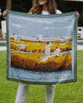 A woman holding up a square silk scarf  featuring a painted landscape with white birds over golden marsh grass under a blue sky.