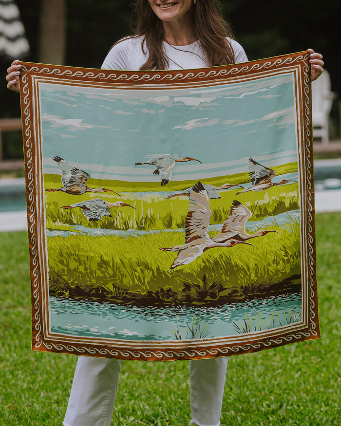 Woman holding a silk scarf with birds flying over a marsh and a decorative border.