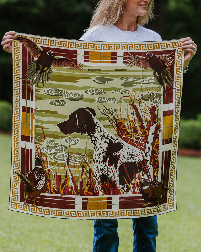 A woman holds up a square silk scarf featuring a German Shorthaired Pointer dog in a marsh with ducks and birds.