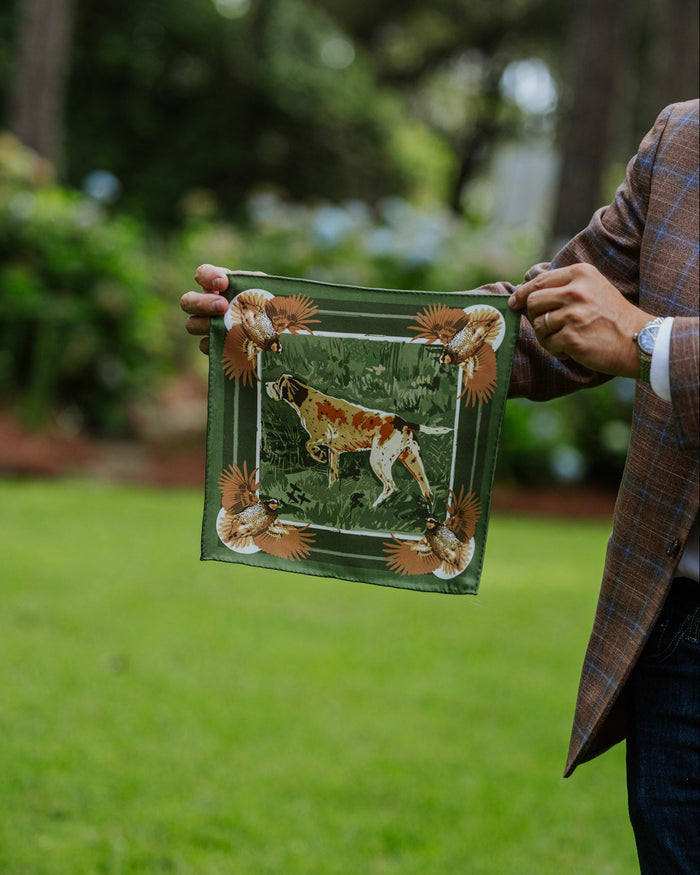 A person outdoors holding a silk pocket square featuring a hunting dog illustration and decorative corners.