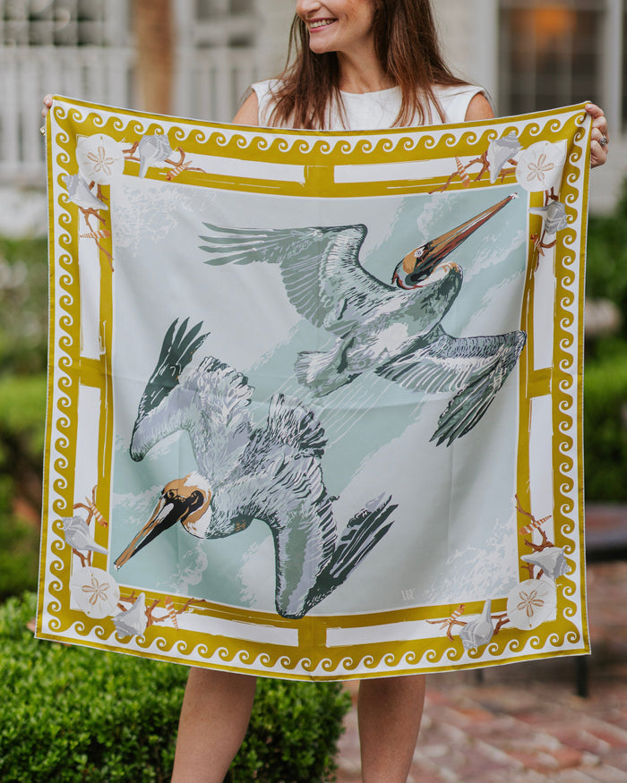 A woman outdoors holds up a square silk scarf featuring two flying pelicans on a pale blue background with a gold and white decorative border.