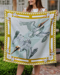 A woman outdoors holds up a square silk scarf featuring two flying pelicans on a pale blue background with a gold and white decorative border.