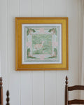 A framed square art print with a gold frame hangs on a white paneled wall above a wooden dining table and chairs.