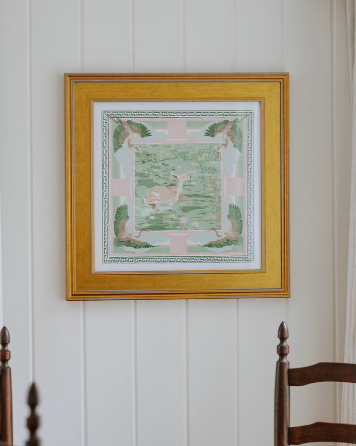 A framed square art print with a gold frame hangs on a white paneled wall above a wooden dining table and chairs.