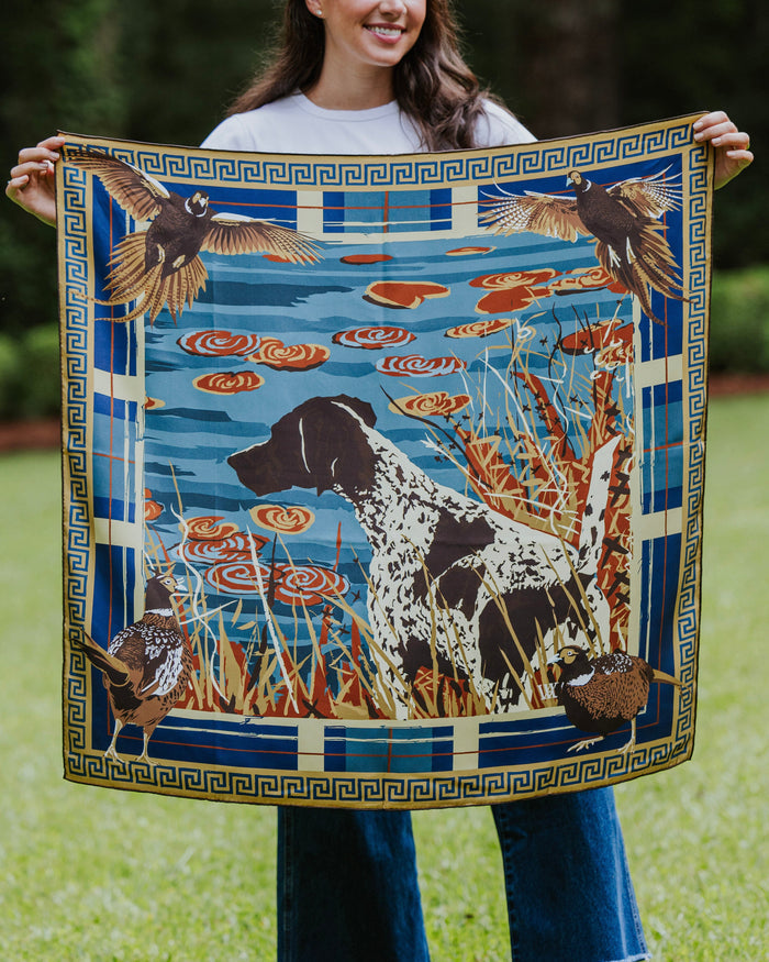 A woman holds up a square silk scarf featuring a hunting dog, birds, lily pads, and a blue border.