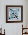 A framed square art print featuring a  dog in a blue-toned hunting scene, hung on a white paneled wall above a wooden dining table and chairs.