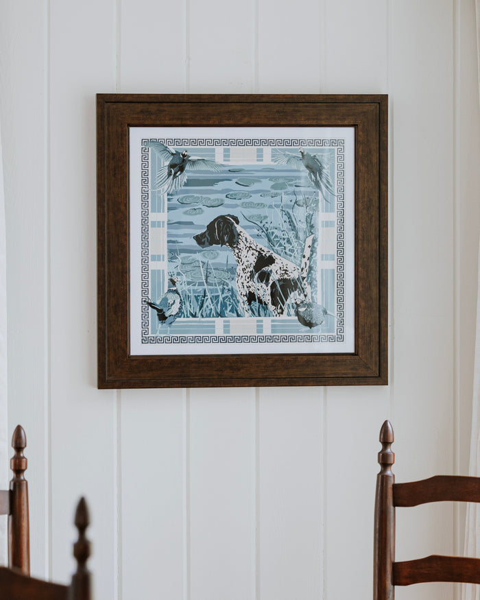 A framed square art print featuring a  dog in a blue-toned hunting scene, hung on a white paneled wall above a wooden dining table and chairs.