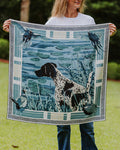 A woman a square silk scarf featuring a blue and white illustration of a black-and-white dog in a marsh with birds and a Greek key border.