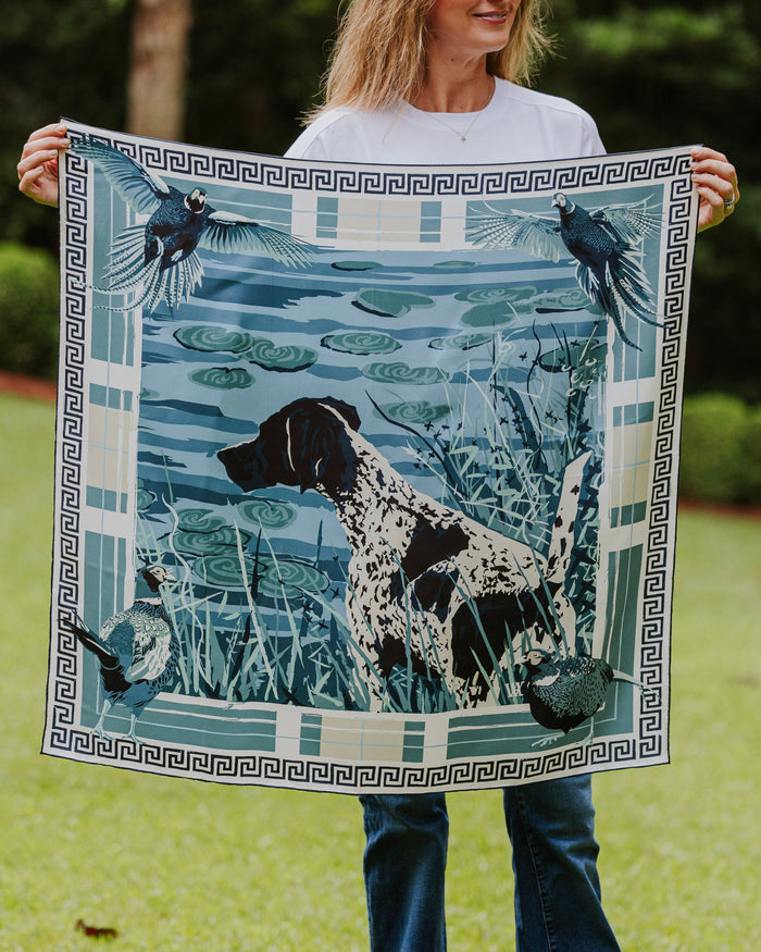 A woman a square silk scarf featuring a blue and white illustration of a black-and-white dog in a marsh with birds and a Greek key border.