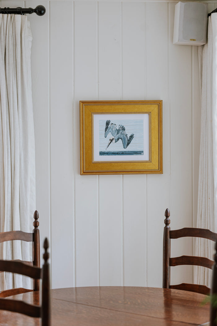 A framed art print depicting a diving pelican in a gold frame, hanging on a white paneled wall above a wooden dining table and chairs.