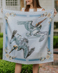 A woman outdoors holds up a square silk scarf featuring two pelicans in flight, with a light blue wave border and shell details.