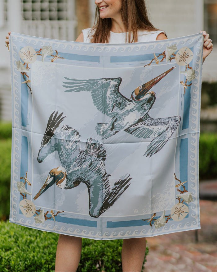A woman outdoors holds up a square silk scarf featuring two pelicans in flight, with a light blue wave border and shell details.