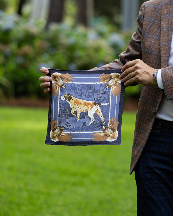 A man holding up a pocket square outdoors on a green lawn. The scarf features a blue background with a hunting dog illustration and ornate gold and brown borders.