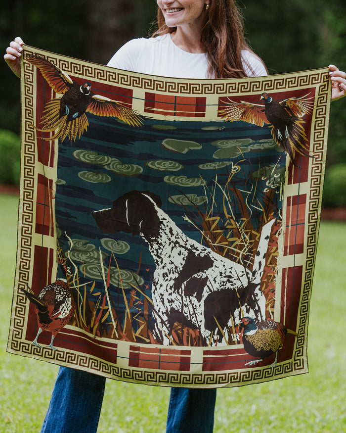 A woman holds up a square silk scarf featuring a hunting dog, birds, and a decorative border.
