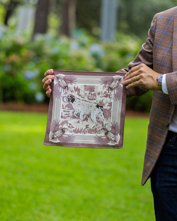 A man in a brown plaid blazer holds up a silk pocket square featuring a hunting dog.