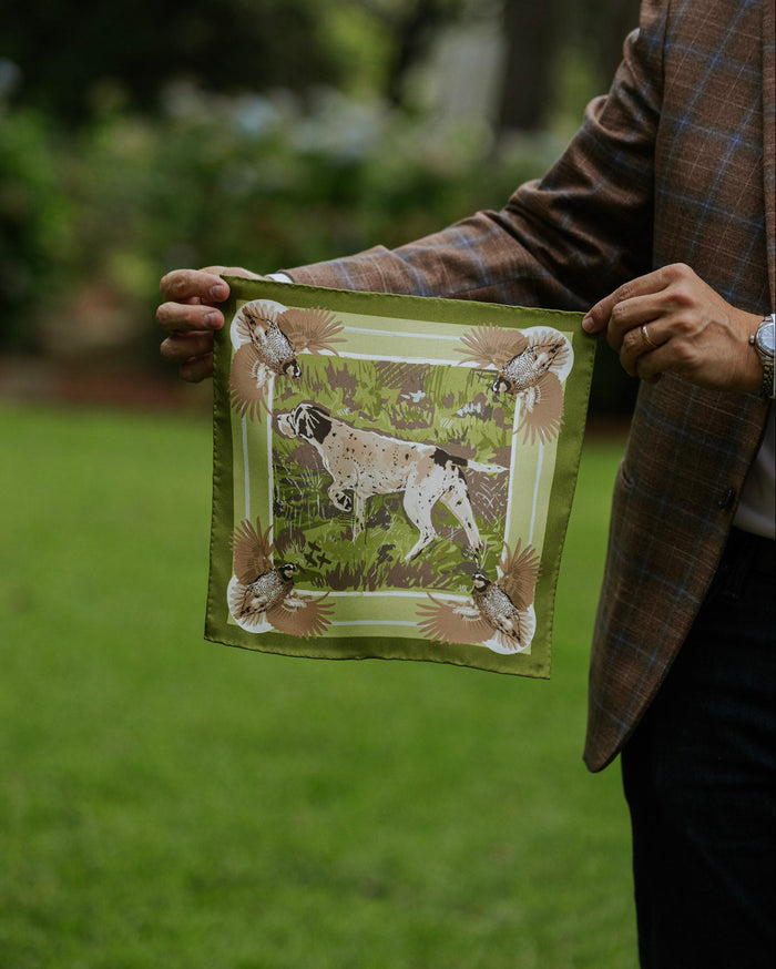 A person in a brown plaid blazer holds up a silk pocket square featuring a hunting dog illustration in a grassy field.