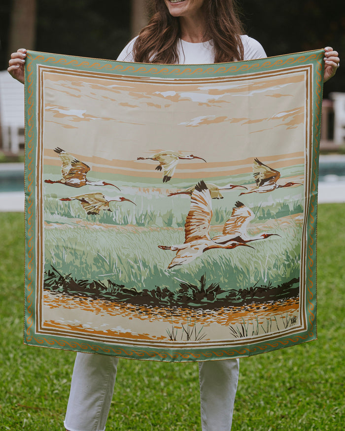 Woman holding a silk scarf with birds flying over a marsh and a decorative border.