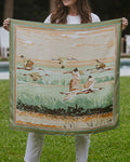 Woman holding a silk scarf with birds flying over a marsh and a decorative border.