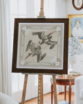 A framed art print featuring two pelicans in flight, displayed on a wooden easel in a bright, modern room with white curtains and a side table.