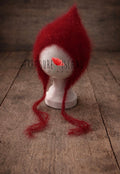 Handmade red newborn pixie bonnet made of soft mohair, displayed on a white mannequin head on a rustic wooden floor background.