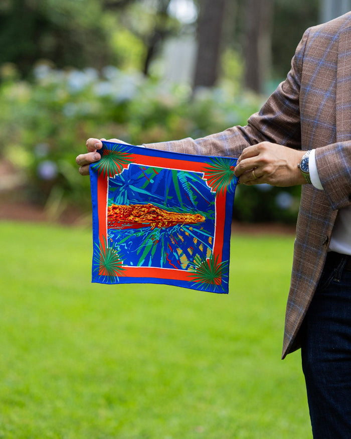 A man in a brown plaid blazer holds up a pocket square featuring a vibrant tropical design with a blue background, palm leaves, and a central red and orange alligator.